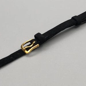 May include: A black watch strap with a gold-colored buckle. The strap is made of a suede-like material and has a rectangular shape. The buckle is a classic design, with a curved shape and a shiny finish.