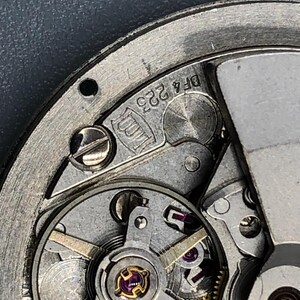 K&ouml;nnte beinhalten: Nahaufnahme eines Uhrwerks mit filigranen Zahnr&auml;dern, Schrauben und einer Unruh. Die Metallkomponenten sind silberfarben, mit Gold- und Lilat&ouml;nen. Der Text "DF4 223" ist auf dem Metall zu sehen.