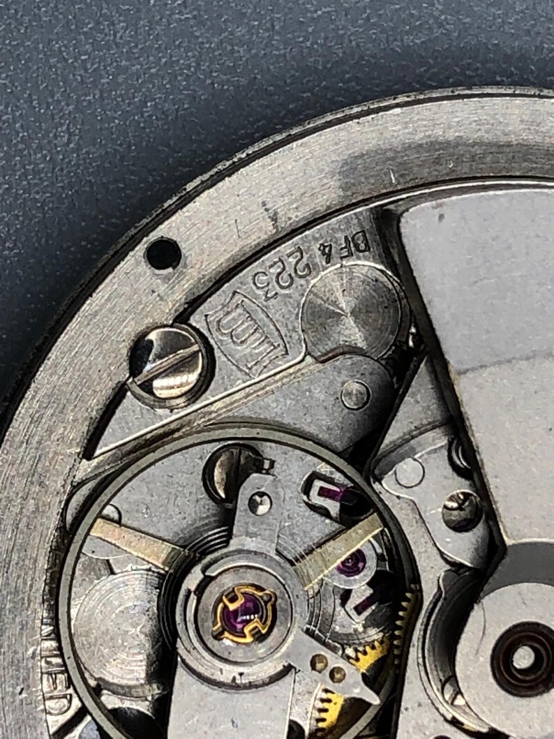 K&ouml;nnte beinhalten: Nahaufnahme eines Uhrwerks mit filigranen Zahnr&auml;dern, Schrauben und einer Unruh. Die Metallkomponenten sind silberfarben, mit Gold- und Lilat&ouml;nen. Der Text "DF4 223" ist auf dem Metall zu sehen.