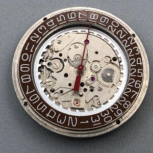 K&ouml;nnte beinhalten: Nahaufnahme eines Uhrwerks mit sichtbaren Zahnr&auml;dern, Juwelen und einem roten Sekundenzeiger. Der &auml;u&szlig;ere Ring zeigt die Tage des Monats in wei&szlig;en Ziffern auf braunem Hintergrund. Detaillierte Uhrwerk-Mechanik.