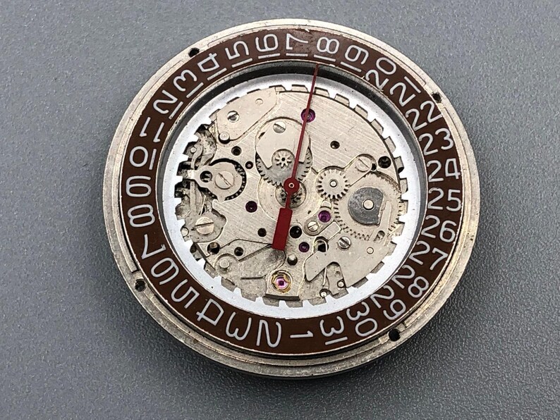 K&ouml;nnte beinhalten: Nahaufnahme eines Uhrwerks mit sichtbaren Zahnr&auml;dern, Juwelen und einem roten Sekundenzeiger. Der &auml;u&szlig;ere Ring zeigt die Tage des Monats in wei&szlig;en Ziffern auf braunem Hintergrund. Detaillierte Uhrwerk-Mechanik.
