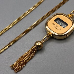May include: A gold-coloured necklace with a pendant watch displaying the text "ASTROSONIC LCD QUARTZ". The watch has a digital display and a tassel. The chain is a delicate, linked design.