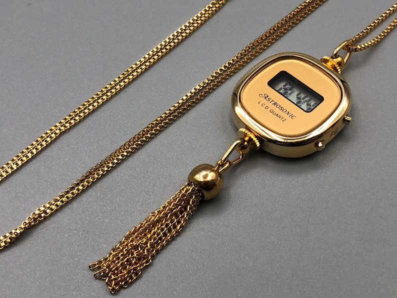 May include: A gold-coloured necklace with a pendant watch displaying the text "ASTROSONIC LCD QUARTZ". The watch has a digital display and a tassel. The chain is a delicate, linked design.