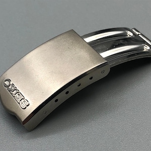 May include: A silver-toned watch clasp with the brand name "SEIKO" embossed on the curved top. The clasp is made of metal and has a curved design with several small holes along the edge.