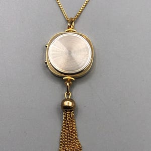 May include: Gold-coloured necklace with a pendant resembling a watch. The pendant has a circular shape with a textured back and a small tassel. The chain and tassel are made of delicate gold links.