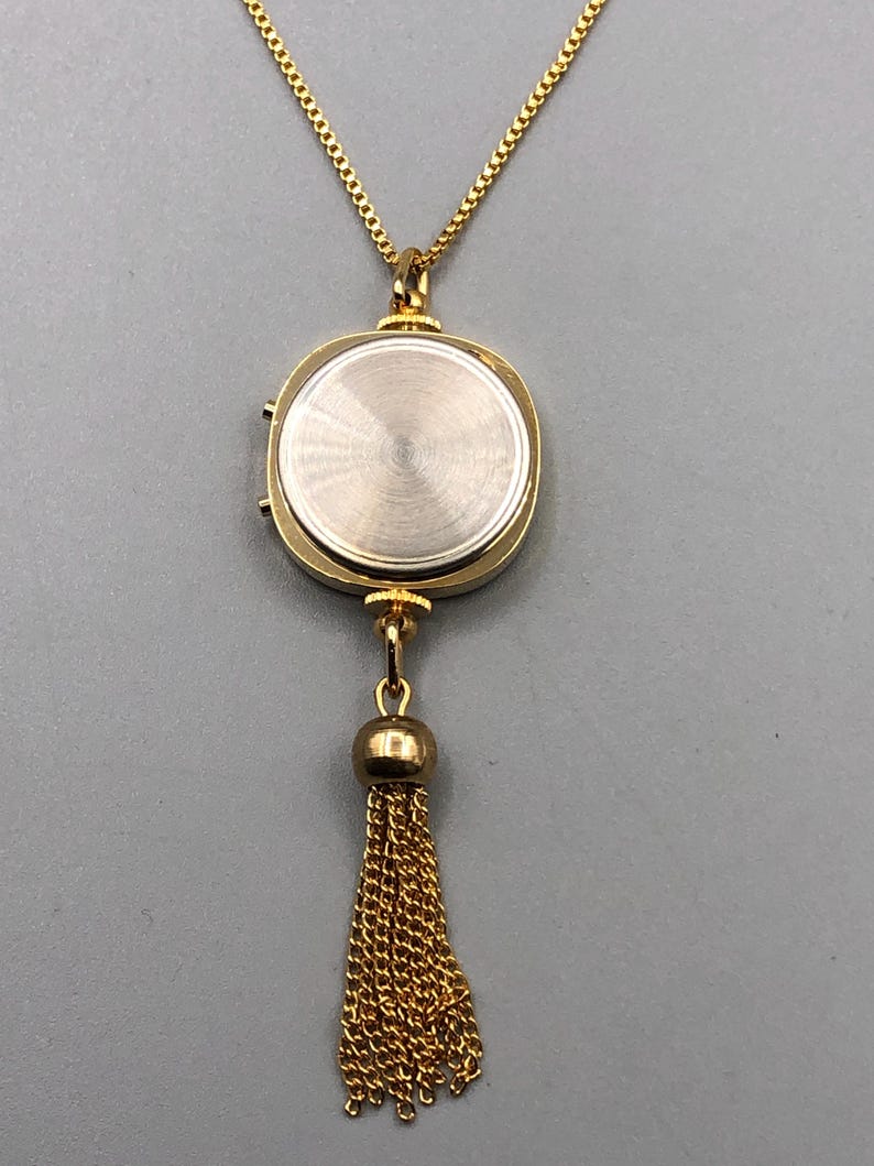 May include: Gold-coloured necklace with a pendant resembling a watch. The pendant has a circular shape with a textured back and a small tassel. The chain and tassel are made of delicate gold links.