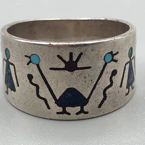 May include: A silver ring with a wide band featuring inlaid designs. The ring has a repeating pattern of stylized figures in blue, green, and black. The ring's design includes geometric shapes and abstract human figures.