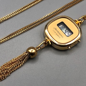 May include: Gold-coloured necklace with a digital watch pendant. The watch displays a digital time and has the text "ASTROSONIC LCD QUARTZ". A gold-coloured chain and tassel are attached to the watch. The tassel hangs below the watch.