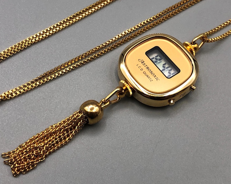 May include: Gold-coloured necklace with a digital watch pendant. The watch displays a digital time and has the text "ASTROSONIC LCD QUARTZ". A gold-coloured chain and tassel are attached to the watch. The tassel hangs below the watch.
