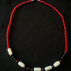 May include: A beaded necklace with a red and black color scheme. The necklace features small red beads and larger, rectangular white beads. The necklace is secured with a silver clasp. The background is black.