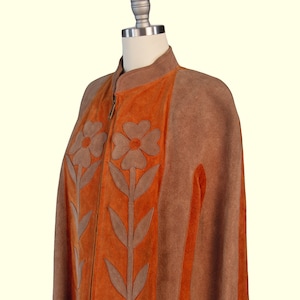 May include: A brown and orange suede cape featuring a high collar, a front zipper closure, and large floral appliqu&eacute; designs in brown suede on the orange front panels. The garment has side arm slits and a structured, sleeveless silhouette.