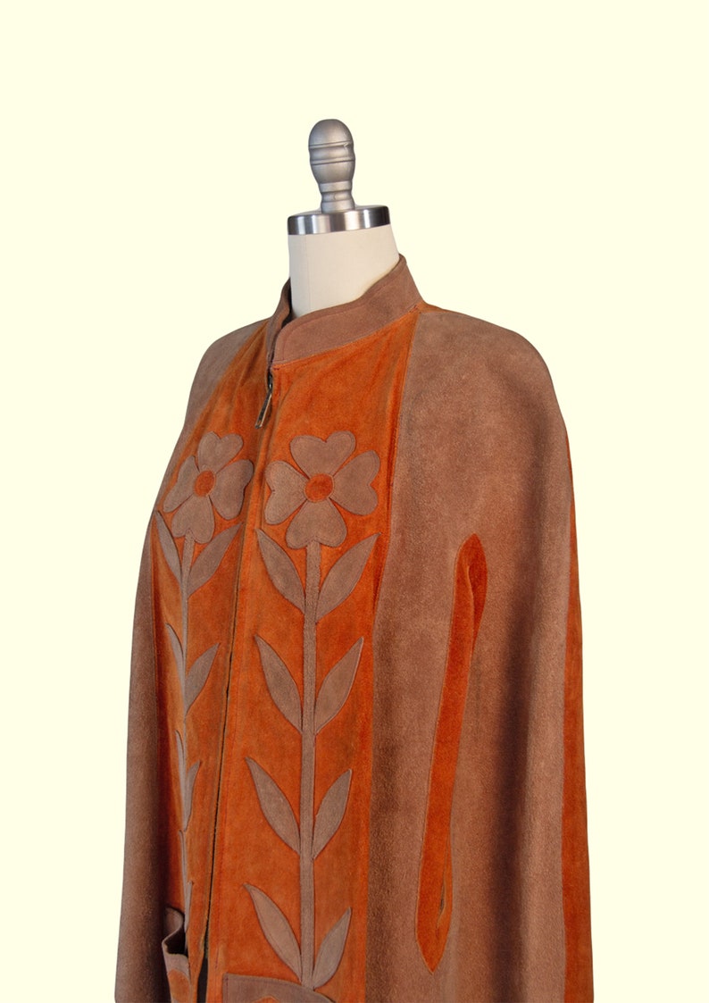 May include: A brown and orange suede cape featuring a high collar, a front zipper closure, and large floral appliqu&eacute; designs in brown suede on the orange front panels. The garment has side arm slits and a structured, sleeveless silhouette.