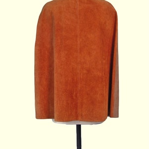 May include: A burnt orange suede cape featuring a smooth tan collar and a vertical seam running down the center back. The garment is displayed on a mannequin, showcasing a sleeveless design with a flared silhouette and a textured finish.
