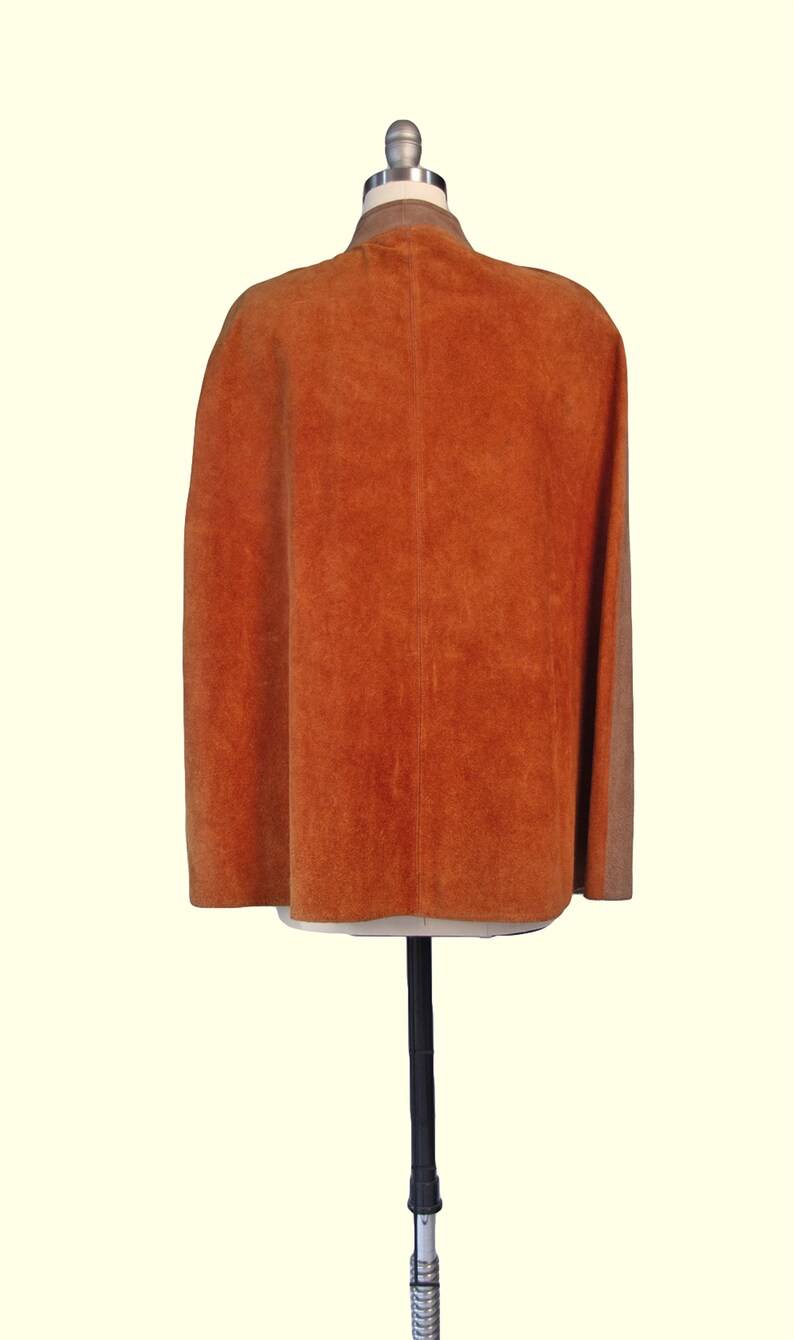 May include: A burnt orange suede cape featuring a smooth tan collar and a vertical seam running down the center back. The garment is displayed on a mannequin, showcasing a sleeveless design with a flared silhouette and a textured finish.