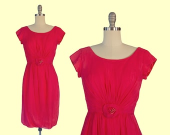 May include: A bright pink vintage dress with short sleeves and a gathered bodice. The dress has a small, jeweled flower embellishment at the waist.