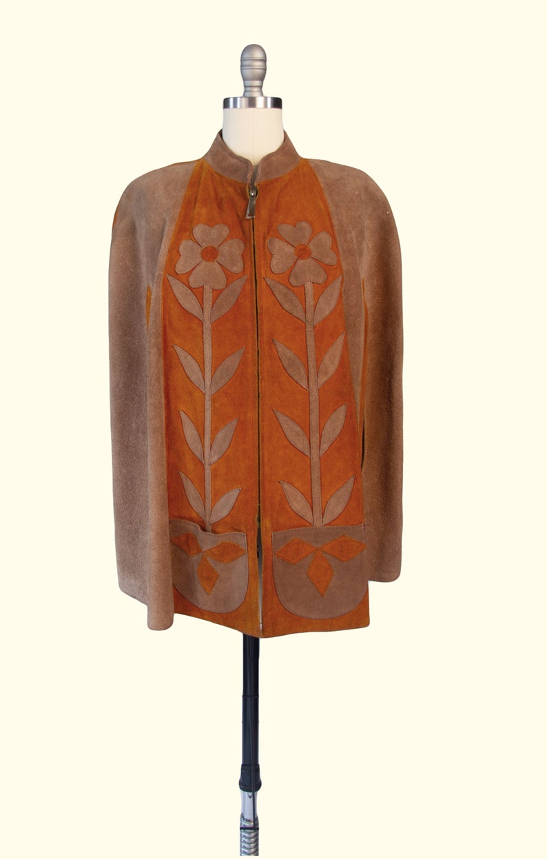 May include: A suede cape in shades of brown and orange featuring a front zipper closure and a high collar. The garment is decorated with large, appliqu&eacute;d floral motifs in brown suede on the orange front panels and two lower patch pockets.