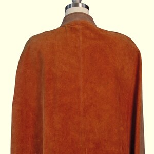 May include: A sleeveless cape made of burnt orange suede leather featuring a central vertical seam down the back and a contrasting light brown collar. The garment is displayed on a mannequin, showcasing the textured surface of the suede material.