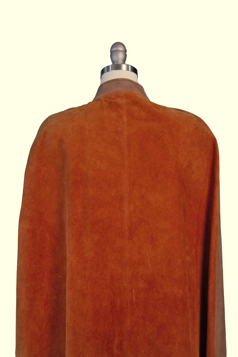 May include: A sleeveless cape made of burnt orange suede leather featuring a central vertical seam down the back and a contrasting light brown collar. The garment is displayed on a mannequin, showcasing the textured surface of the suede material.