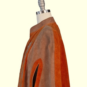 May include: A suede cape featuring alternating vertical panels of tan and burnt orange. The garment has a high collar, a front closure, and arm slits. The texture of the suede is visible throughout the piece, showcasing the distinct color blocking.