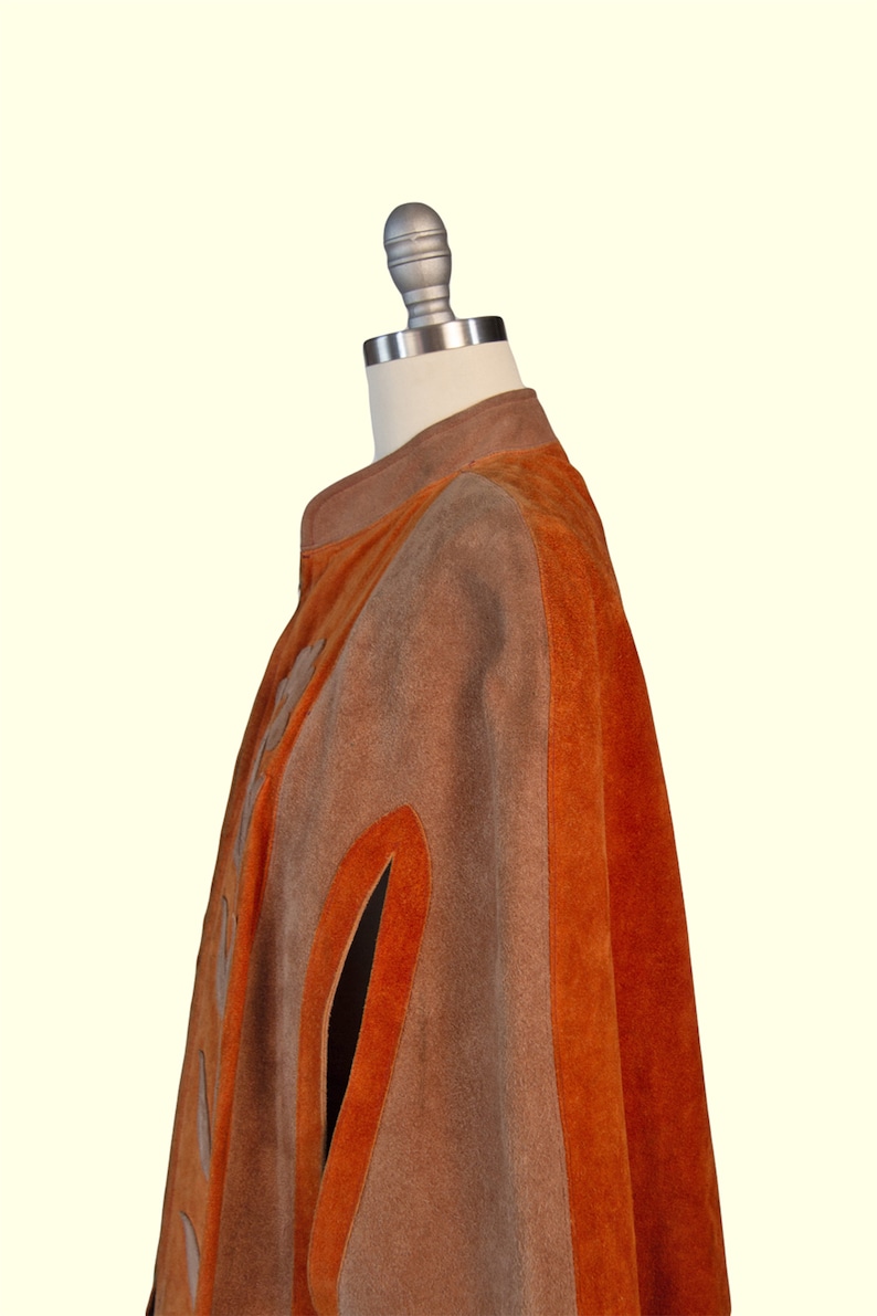 May include: A suede cape featuring alternating vertical panels of tan and burnt orange. The garment has a high collar, a front closure, and arm slits. The texture of the suede is visible throughout the piece, showcasing the distinct color blocking.
