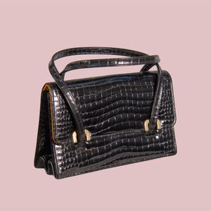 May include: A black handbag with a glossy crocodile-embossed texture. The bag features a structured rectangular shape, a single top handle, and gold-toned metal hardware accents where the handle attaches to the front flap of the bag.
