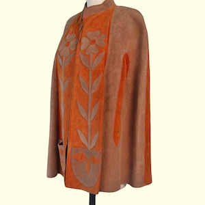 May include: A brown and orange suede cape featuring a front zipper closure, a high collar, and two front pockets. The garment is decorated with large, light brown floral and leaf applique designs that extend vertically along the front panels.