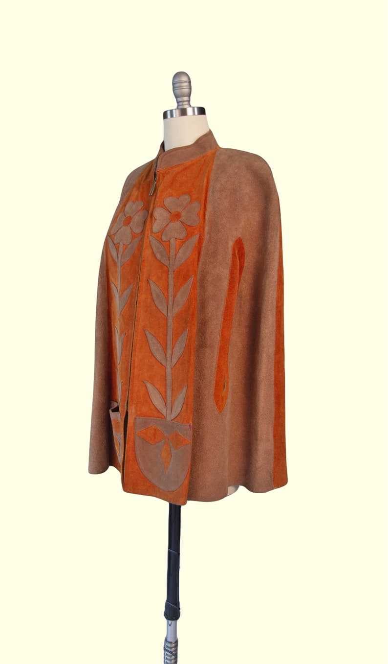 May include: A brown and orange suede cape featuring a front zipper closure, a high collar, and two front pockets. The garment is decorated with large, light brown floral and leaf applique designs that extend vertically along the front panels.