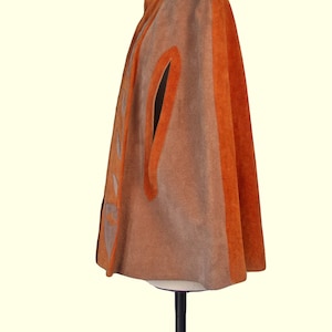 May include: A suede cape in shades of tan and burnt orange, featuring a high collar, arm slits, and a vertical panel along the front closure decorated with a series of triangular cutouts. The garment is displayed on a mannequin stand.