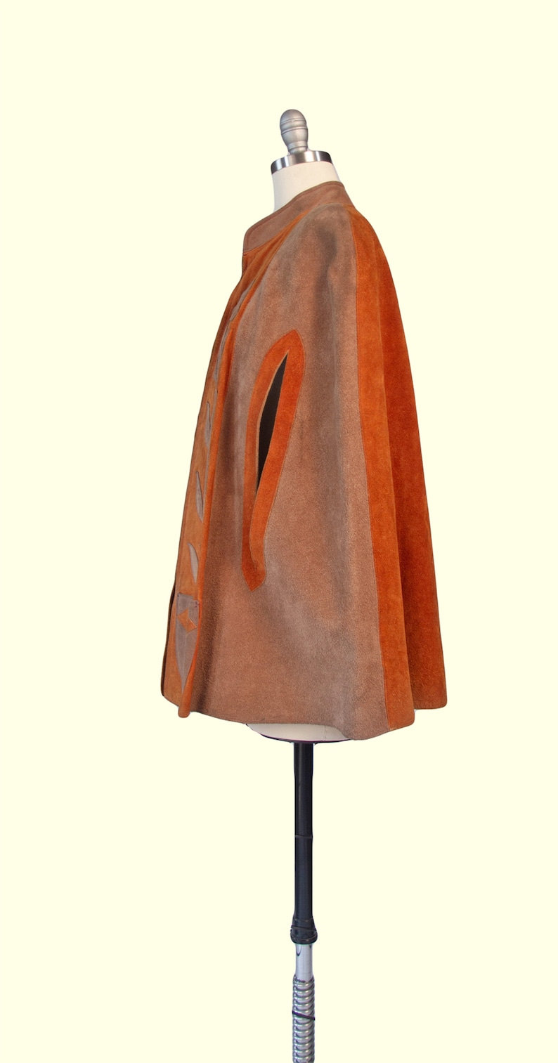 May include: A suede cape in shades of tan and burnt orange, featuring a high collar, arm slits, and a vertical panel along the front closure decorated with a series of triangular cutouts. The garment is displayed on a mannequin stand.