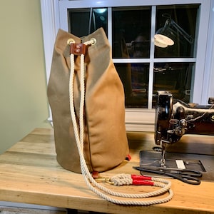 Può includere: Una borsa a sacco in tela color cachi con chiusura a coulisse e dettagli in pelle. La borsa cilindrica è in posizione verticale. Una corda abbinata con dettagli rossi è davanti. Sullo sfondo, una macchina da cucire Singer vintage.