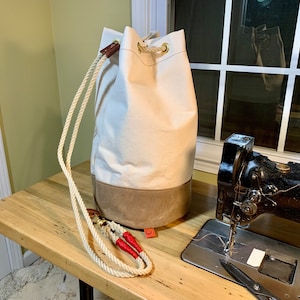 May include: A white canvas duffel bag with brown leather trim and a rope handle. The bag is sitting on a wooden table next to a black sewing machine.
