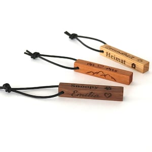 Può includere: Tre portachiavi in legno con cordini neri. Il portachiavi superiore è di colore marrone chiaro con il testo "Gemütlichkeit Heimat" e un'icona di casa. Il portachiavi centrale è di colore marrone con il testo "1913 ♥ 2013" e un'icona di montagna. Il portachiavi inferiore è di colore marrone scuro con il testo "Snoopy Emilio" e un'icona di impronta di zampa e cuore.