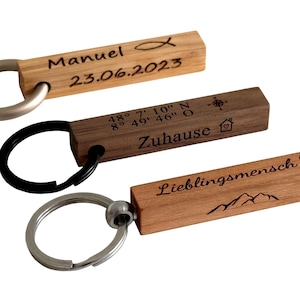 Op de afbeelding: Drie houten sleutelhangers met metalen ringen. Eén sleutelhanger heeft de naam "Manuel" en de datum "23.06.2023" gegraveerd. Een andere heeft coördinaten en het woord "Zuhause". De laatste zegt "Lieblingsmensch".