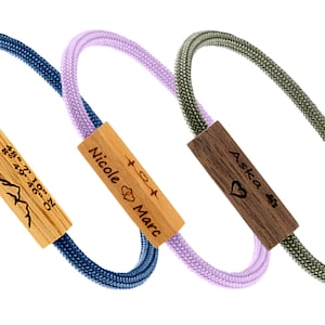 Può includere: Tre braccialetti personalizzati con perline di legno. Il braccialetto blu ha le coordinate incise sulla perlina, il braccialetto viola ha i nomi "Nicole" e "Marc" incisi sulla perlina, e il braccialetto verde ha il nome "Aska" e un cuore inciso sulla perlina.