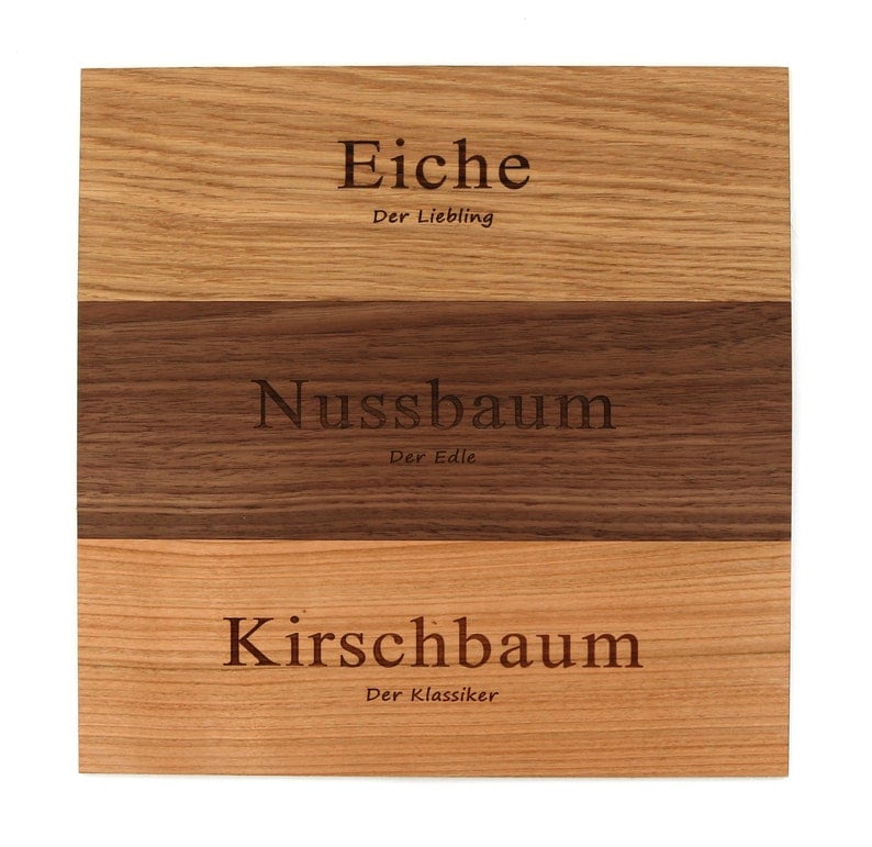 May include: Three wooden boards with different wood grains. The top board is light brown and says "Eiche Der Liebling". The middle board is dark brown and says "Nussbaum Der Edle". The bottom board is light brown and says "Kirschbaum Der Klassiker".