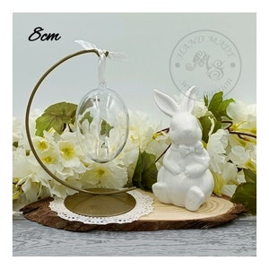 May include: A white ceramic bunny figurine sits on a wooden slice with a gold metal stand holding a clear glass egg ornament. The stand is 8 cm tall. The bunny has a bow tie and is sitting upright. The wooden slice is surrounded by white flowers.
