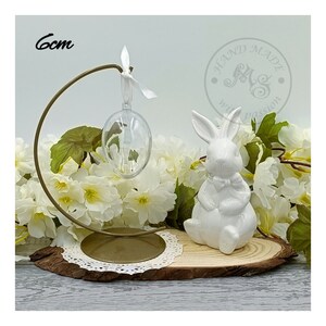Pode incluir: Uma figurinha de coelho de cerâmica branca está sentada em uma fatia de madeira com um suporte de metal dourado que segura um ornamento de ovo de vidro transparente. O suporte é decorado com uma renda branca. O coelho tem uma gravata borboleta e mede 6 cm de altura.