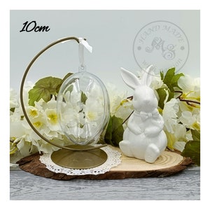 May include: A clear glass egg ornament with a white metal stand, a white ceramic bunny figurine, and a wooden base with a white lace doily. The ornament is 10cm in diameter.