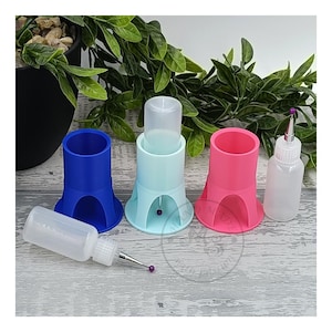 Könnte beinhalten: Drei Flaschenhalter in Blau, Hellblau und Rosa, jeweils mit einer durchsichtigen Flasche darin. Eine durchsichtige Flasche mit einem kleinen Auslauf befindet sich rechts im Bild. Eine durchsichtige Flasche mit einem kleinen Auslauf und einer lila Perle befinden sich links im Bild.