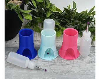 May include: Three blue, light blue, and pink bottle holders with a clear bottle in each holder. A clear bottle with a small nozzle is on the right side of the image. A clear bottle with a small nozzle and a purple bead are on the left side of the image.