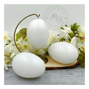May include: Three white styrofoam eggs on a wooden stand with a metal stand behind them. The eggs are smooth and round. The stand is brown and has a natural wood finish. The metal stand is gold and has a curved design. The background is white with a few white flowers.