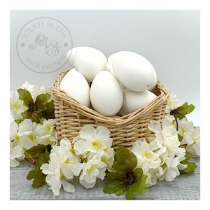Op de afbeelding: Een rieten mand gevuld met wit geverfde eieren, omringd door witte bloemen met groene bladeren. De mand heeft een label met de tekst "Hand Made with passion".
