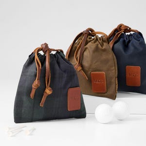 May include: Three golf ball pouches in navy, brown, and dark green, each with a leather patch displaying "MKG". The pouches have leather drawstring closures and are accompanied by two golf balls and golf tees.
