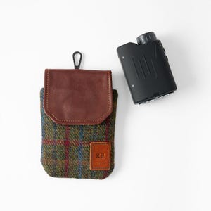 May include: A black rangefinder and a green, blue, and red plaid tweed case with a brown leather flap and a small leather patch with the initials "RB". The case has a black carabiner clip.