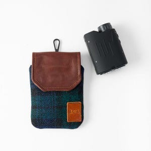 May include: A black rangefinder and a case. The case is made of a blue and green plaid fabric with a brown leather flap and a small leather patch with the initials "LCR". The rangefinder is black.