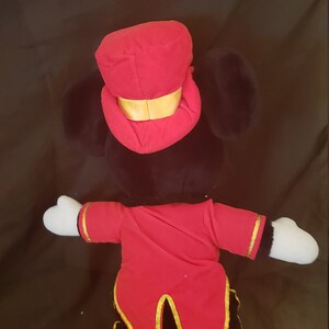 May include: A plush toy of Mickey Mouse dressed as a ringmaster. He is wearing a red and gold costume with a black top hat. He has black ears and a white face.