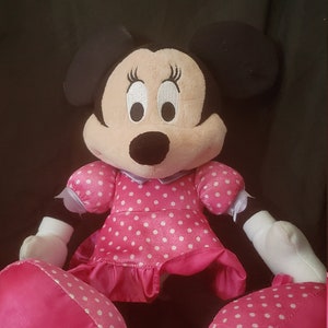 Pu&ograve; includere: Un vestito a pois rosa e bianchi con un fiocco di raso rosa. Il giocattolo di peluche &egrave; un personaggio dei cartoni animati con orecchie nere e occhi grandi.