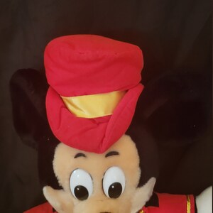 May include: A plush Mickey Mouse toy wearing a red and gold conductor's uniform. The toy has large black eyes and a black nose.