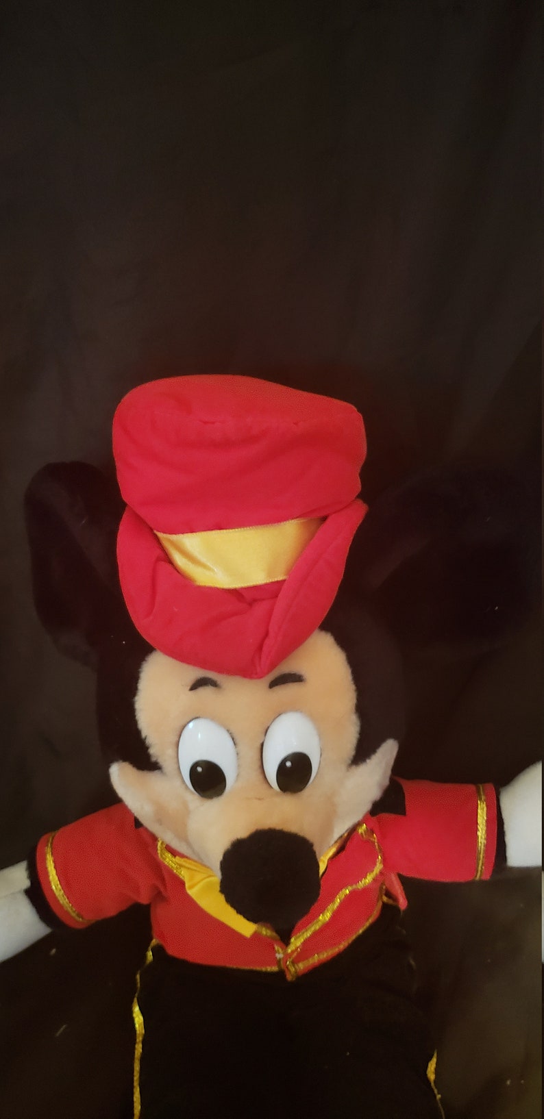 May include: A plush Mickey Mouse toy wearing a red and gold conductor's uniform. The toy has large black eyes and a black nose.