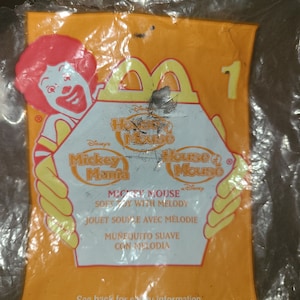 May include: A yellow plastic bag with a Mickey Mouse Mania toy inside. The bag features a red and yellow Mickey Mouse character and the text "Mickey Mouse Mania" and "House of Mouse".